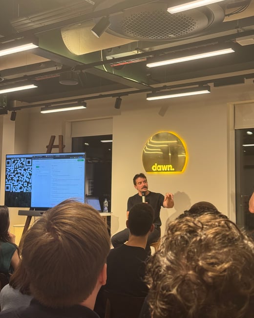 A photograph taken from the audience perspective capturing a man giving a seated presentation or talk in a modern event space, with a large screen displaying a QR code and an illuminated 'dawn.' logo visible behind him.