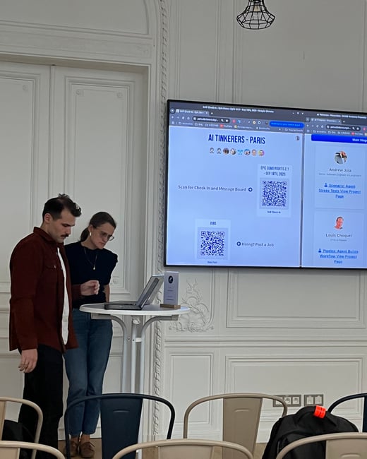 A candid photograph taken indoors showing two individuals standing near a small table with an open laptop, while a large screen behind them displays information for an event titled 'AI Tinkerers - Paris'. The setting is an elegant room characterized by white, ornate wall paneling.