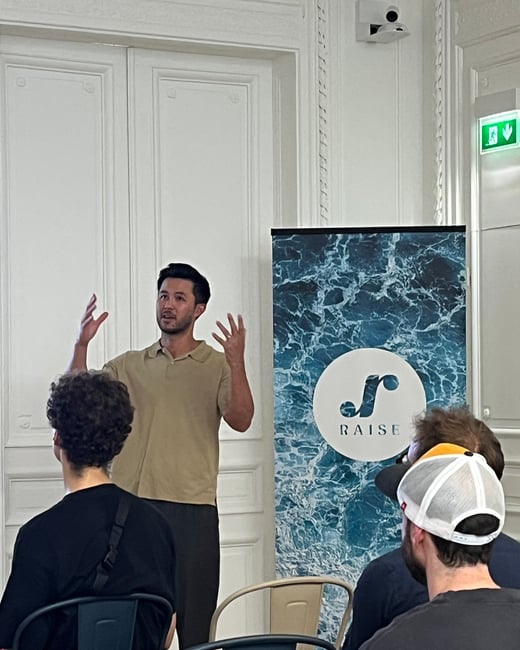 A photograph capturing a man giving a presentation or talk indoors, standing next to a vertical banner for 'RAISE' and addressing several audience members seated in the foreground.
