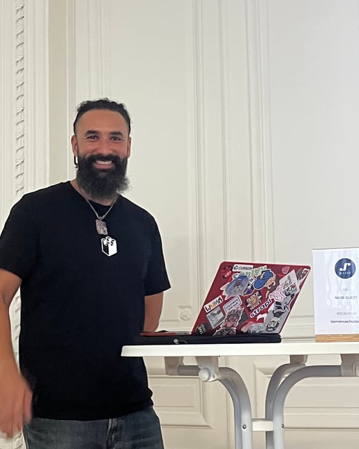 A candid photo of a smiling man with a dark beard and black t-shirt, standing next to a small table holding a sticker-covered red laptop and a small informational sign, likely at an indoor event.