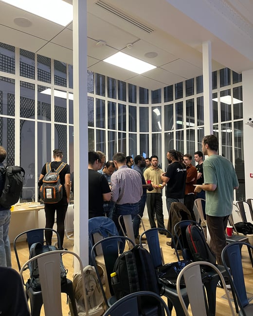 A candid photograph capturing a group of approximately 18 people socializing and networking indoors, likely during a break or after an event, in a modern space with large, multi-paned windows.