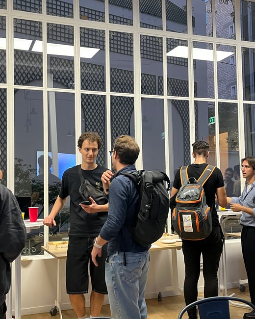 A candid photograph capturing a group of people socializing and talking indoors, set against a distinctive large glass wall with white framing and black decorative lattice work.