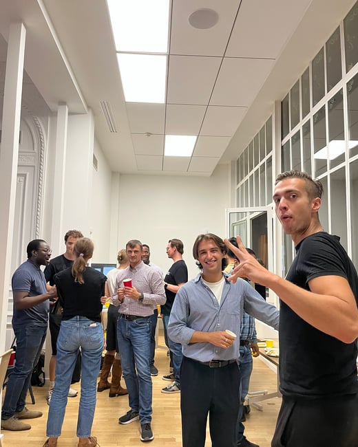 A candid photograph capturing a group of approximately a dozen people socializing and holding drinks in a brightly lit, modern indoor hallway or office space, with two men posing in the foreground.