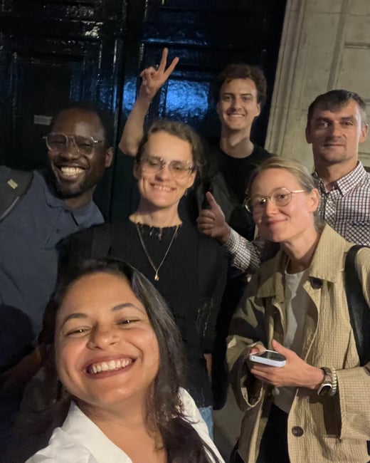 A low-light group selfie captures six smiling people posing closely together against a dark background, with several individuals making hand gestures like a peace sign and a thumbs up.