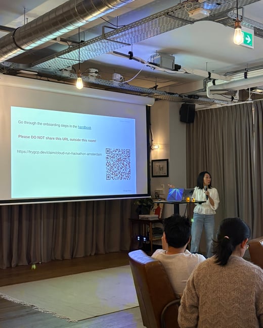 A woman stands in an indoor space giving a presentation, pointing at a large projection screen displaying onboarding instructions and a QR code.