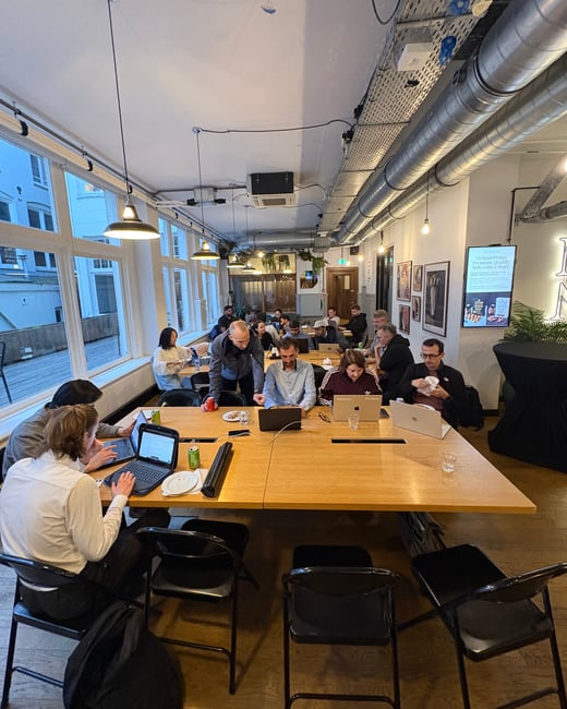 A group of people are working on their laptops in a modern, industrial-style coworking space or office with exposed ceiling pipes and large windows.