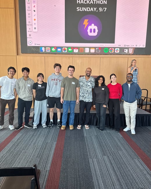 A group of nine people, likely participants and organizers of a tech event, are standing together for a photo in front of a large screen displaying information for a hackathon.