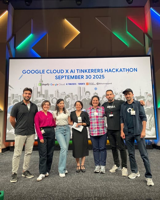A group of seven individuals poses for a photo on a stage beneath a large screen advertising the "Google Cloud X AI Tinkerers Hackathon."