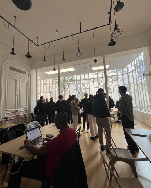 A candid photograph taken indoors showing a large, high-ceilinged room where a man works on a laptop at a table in the foreground, while a group of people stand and socialize near a wall of bright windows in the background.