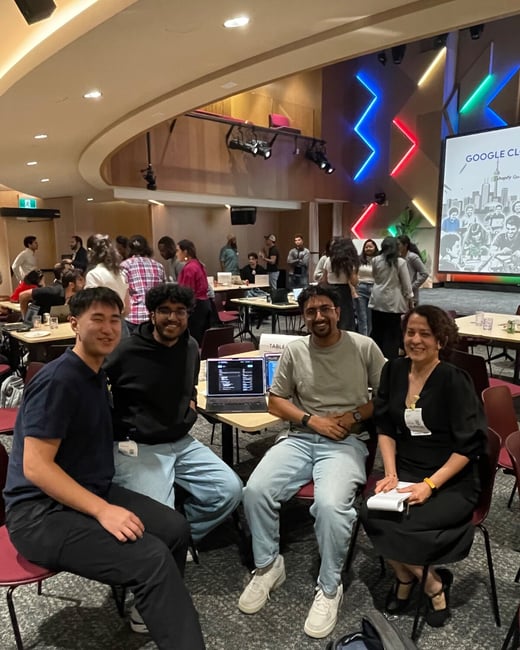 A group of four people, three men and one woman, are seated and smiling directly at the camera at an indoor tech event or conference.