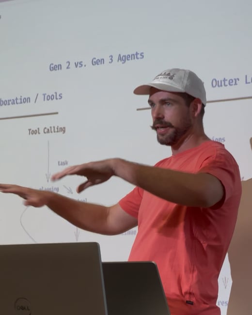 A man with a prominent mustache and beard, wearing a coral t-shirt and a white baseball cap, is giving a presentation while gesturing with both hands in front of a projected screen.