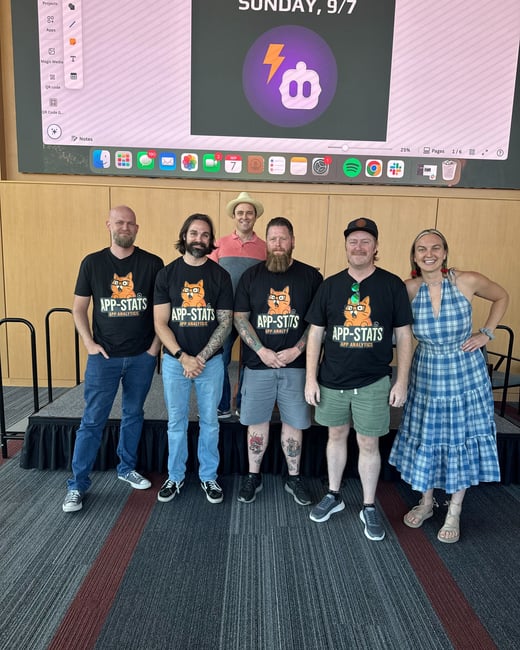 A group photo of six people, likely a development or analytics team, standing in front of a large presentation screen at an indoor event or conference. Four members are wearing matching black t-shirts featuring the 'APP-STATS APP ANALYTICS' logo.