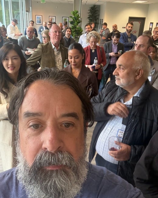 A man with a grey beard takes a selfie in the foreground, with a large crowd of people gathered in an indoor office or event space behind him.