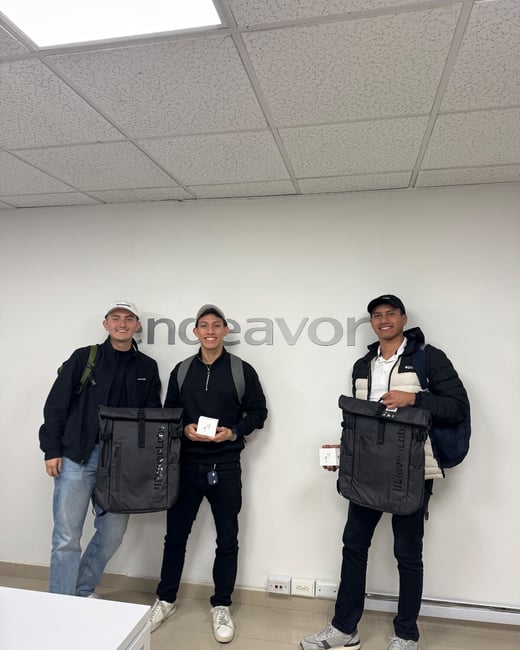 Three young men stand indoors in front of a white wall displaying the large gray word "endeavor," posing with matching black backpacks and small white boxes.