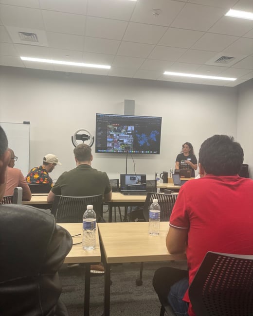 A candid photograph taken from the perspective of an audience member showing a woman presenting information displayed on a large monitor in a modern classroom or workshop setting.