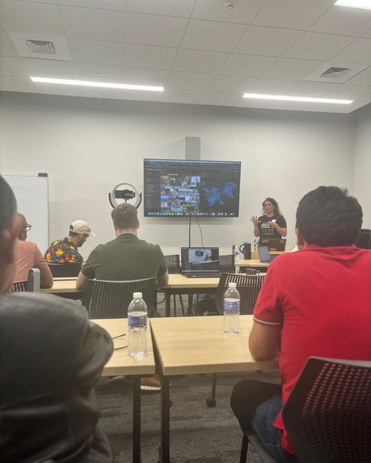 A photograph taken from the audience perspective showing a presenter, identified as a woman, standing in front of a large screen displaying complex visual data during an indoor presentation or workshop.
