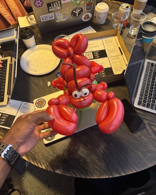 A person's hand holds a detailed red balloon animal shaped like a lobster over a table cluttered with laptops and coffee cups.
