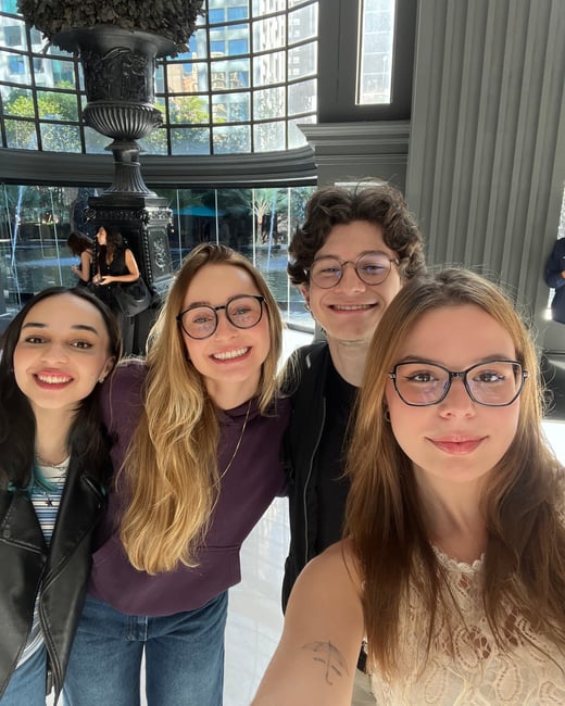 A group of four smiling young adults, three women and one man, posing for a selfie in a bright, modern indoor space with a large decorative fountain in the background.
