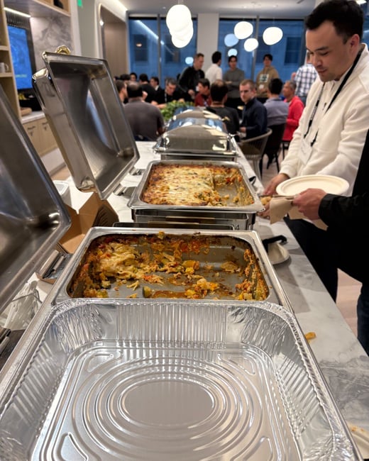 A candid photograph captures a buffet line set up in a modern office or event space, showing chafing dishes containing food, some of which is nearly finished, as a man in the foreground serves himself a plate.
