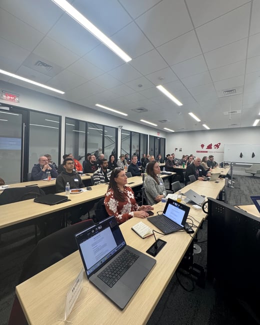 A wide-angle photograph capturing an audience of approximately thirty people seated at long tables in a modern conference room, attending a presentation or meeting. Several laptops and notebooks are visible on the desks in the foreground.