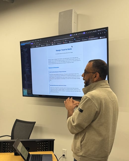 A photograph captures a man standing indoors, facing a large wall-mounted monitor that displays a webpage titled 'Raleigh: Travel for Nerds,' while gesturing with his hands. A laptop is open on a black and wood desk in the foreground.