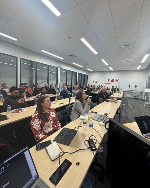 A wide-angle photograph capturing a group of approximately 35 people seated at long tables in a modern conference room, attending a presentation or meeting.
