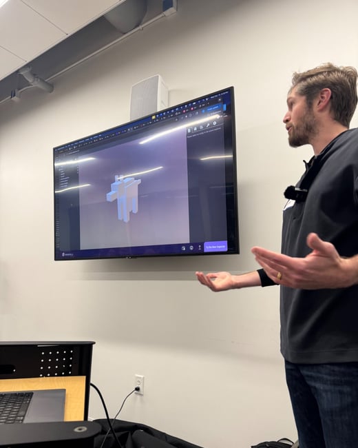 A man stands next to a large wall-mounted monitor, gesturing with open hands while giving a presentation or demonstration of 3D modeling software in an indoor office or conference room setting.