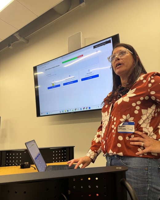 A woman wearing a rust-colored floral shirt and glasses is standing in a classroom or meeting room, presenting content displayed on a large wall-mounted screen labeled "Data Science Project Grader."