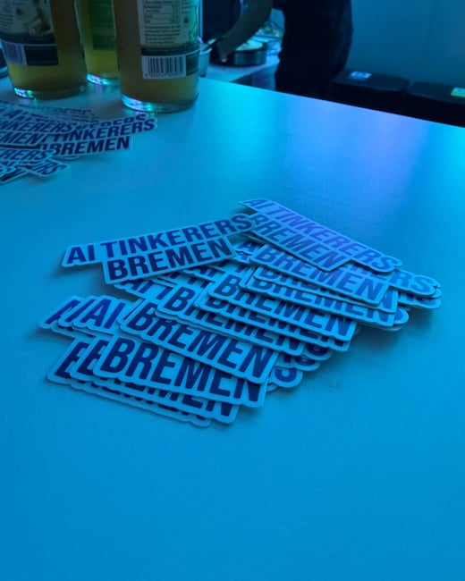 A close-up photograph of several stickers with the text 'AI TINKERERS BREMEN' scattered on a table under intense blue lighting.