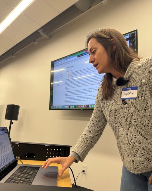 A candid photograph shows a woman named Beatriz, wearing a gray knit sweater, leaning over a laptop while standing near a large monitor displaying a text document, likely in a meeting or presentation setting.