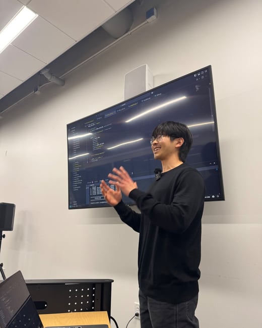 A young man wearing glasses and a black sweater stands gesturing with his hands while giving a technical presentation in front of a large monitor displaying code.
