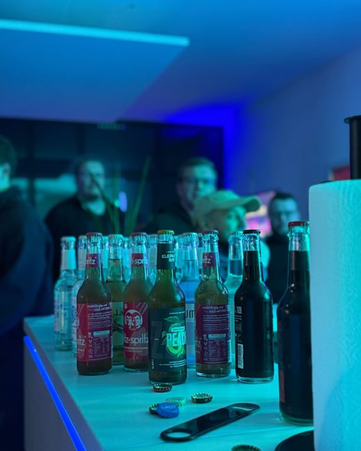 A row of bottled beverages, including Fritz-spritz and Elephant Bay, sits on a counter illuminated by vibrant blue lighting with several people blurred in the background.