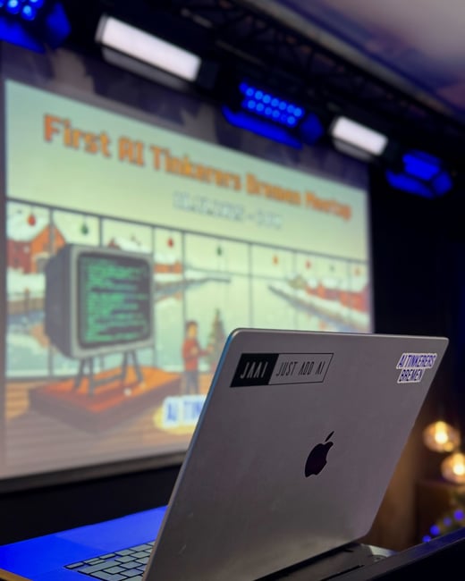 A close-up photograph of a silver laptop with stickers, positioned in front of a projection screen displaying a presentation for the 'First AI Tinkerers Bremen Meetup'.