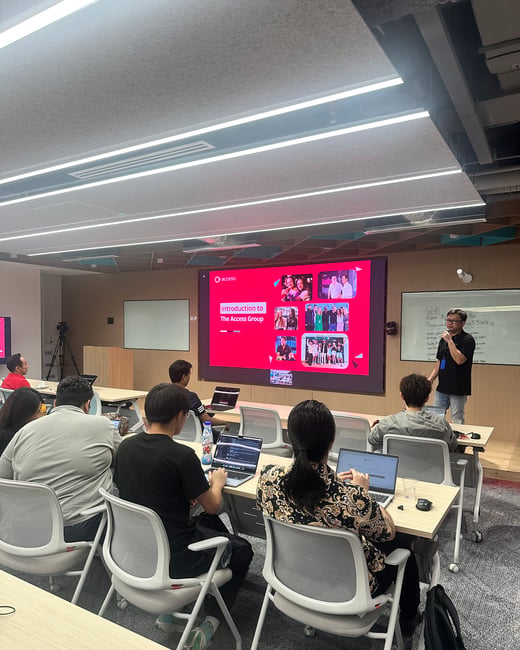 A photograph captures an indoor training session or presentation where several attendees are seated at desks with open laptops, facing a large screen displaying a presentation slide with a red background about 'The Access Group.' A male presenter stands to the right, addressing the audience.