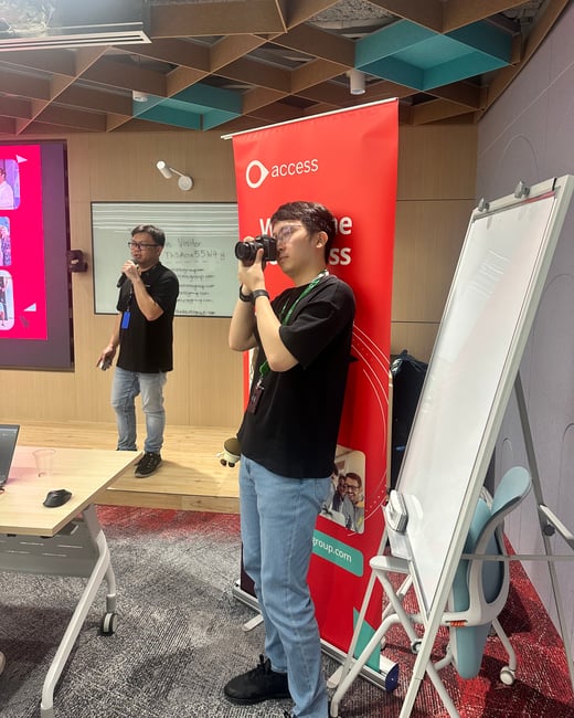 A candid photograph taken indoors during a corporate event, showing two men: one speaking into a microphone during a presentation, and another actively documenting the event by taking photos with a camera in front of a large red banner featuring the 'access' logo.