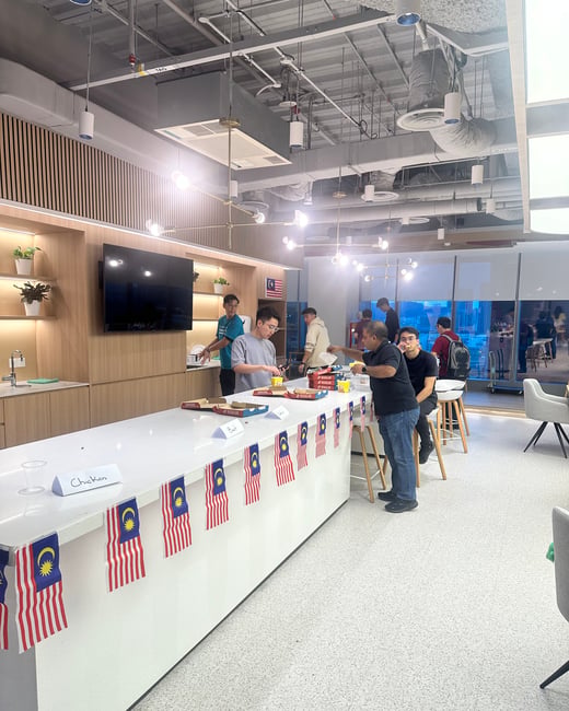 A candid photo capturing a group of people socializing and serving themselves food (likely pizza) at a long counter in a modern office breakroom, which is decorated with a garland of small Malaysian flags.