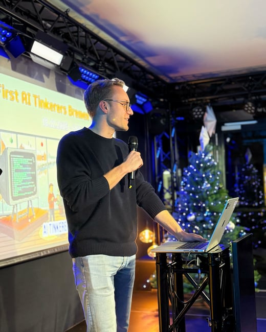 A man is giving a presentation on a stage, holding a microphone and standing next to a laptop with a projector screen in the background.