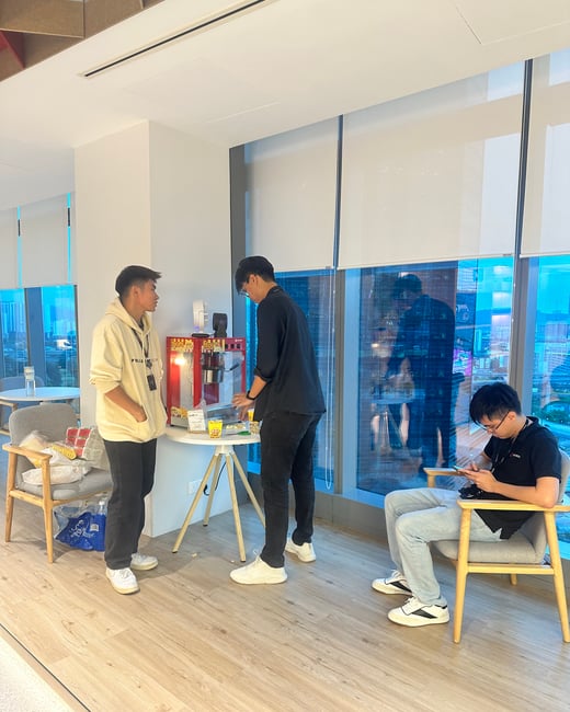 A photograph capturing three young men gathered around a small table with a red popcorn machine in a modern office lounge area. Two men are standing near the machine, while the third is seated nearby looking at his phone, set against a backdrop of large windows showing a blue-hued city view.