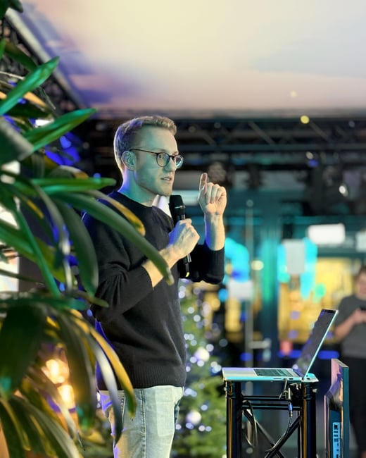 A man wearing glasses and a dark sweater is giving a presentation, holding a microphone and gesturing with one hand while standing next to a laptop.