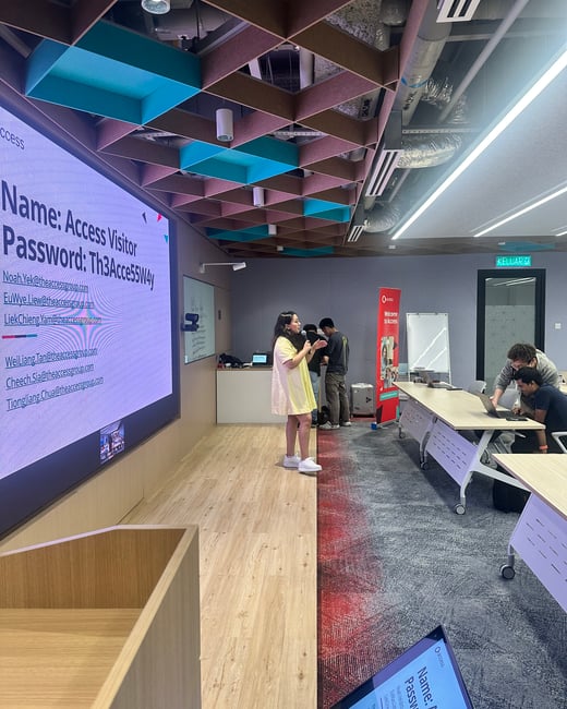 A photograph capturing an indoor presentation or meeting in a modern office space, featuring a large screen displaying Wi-Fi access details and several people engaged in activities, including one person standing near the screen, potentially giving a presentation.