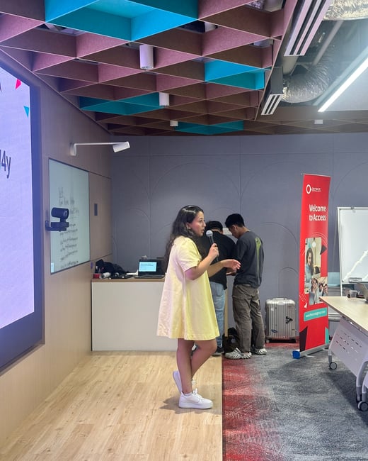 A woman in a light yellow dress is giving a presentation or talk, holding a microphone in a modern indoor setting with a geometrically patterned, multi-colored acoustic ceiling.