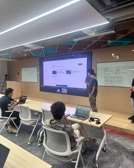 A photograph taken indoors showing a small group of people attending a presentation or workshop in a modern meeting room. One person stands speaking near a large screen displaying a web application, while two attendees sit at desks with open laptops.