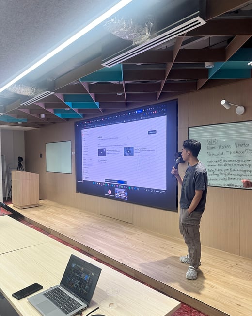 A young man is giving a presentation using a microphone in a modern conference room, standing on a wooden stage in front of a very large display screen showing a web interface. A laptop, headphones, and a smartphone are visible on a table in the foreground.