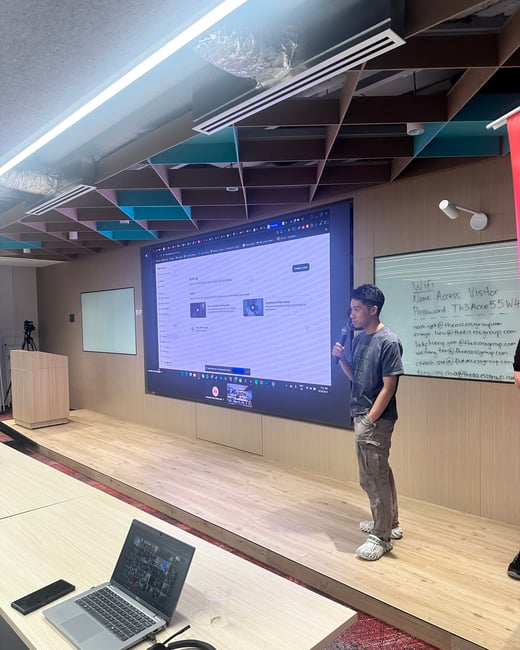 A photograph capturing a man giving a presentation or training session in a modern conference room, standing on a small stage in front of a large screen displaying software content.