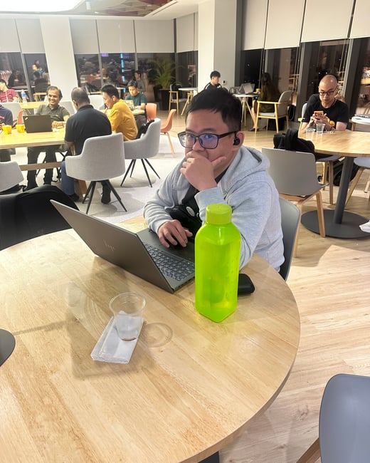 A man wearing glasses and a grey hoodie works on a laptop at a wooden round table, next to a bright neon green water bottle, in a modern, well-lit communal office or co-working space during the evening.