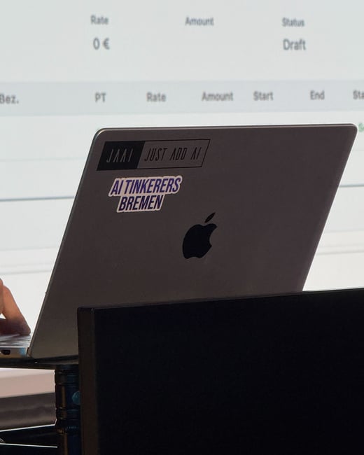 A silver Apple laptop with stickers on its lid is positioned in front of a screen displaying a table of data.