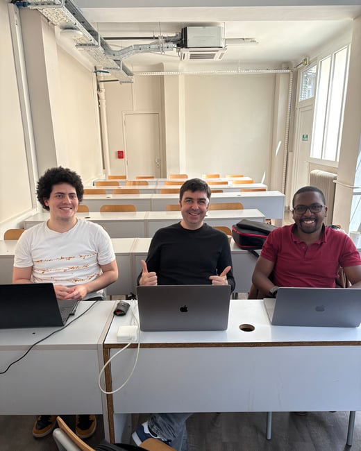 Three men are sitting at a long white desk in a classroom setting, each with a laptop open in front of them. The man in the center is smiling and giving a double thumbs-up gesture.