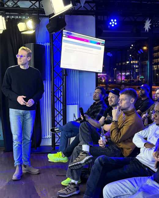 A man is giving a presentation in a room illuminated with blue light, while an audience of several people listens. A screen behind him displays text about software version releases and pricing plans.
