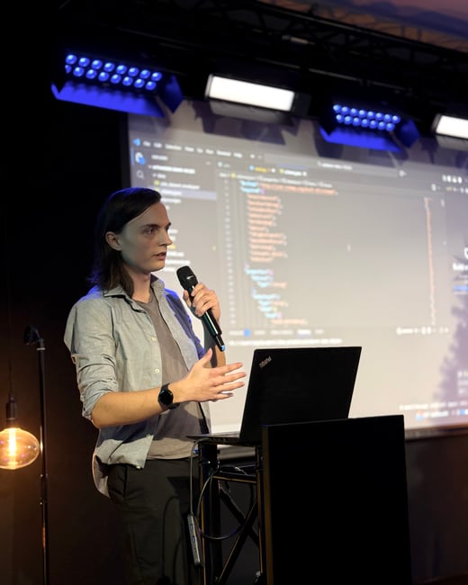 A person with long hair is giving a technical presentation on a stage, holding a microphone and gesturing while a screen behind them displays a code editor.