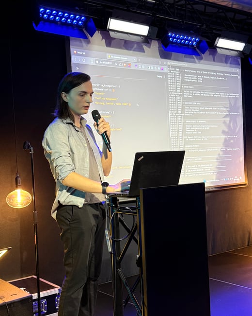 A person stands on a stage giving a presentation, holding a microphone in one hand and using a laptop with the other. A large screen behind them displays code and log data related to a job metadata generator.